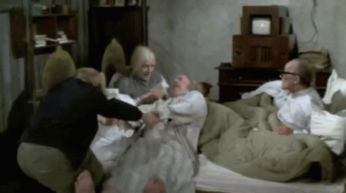 A GIF from the movie Willy Wonka & the Chocolate Factory showing the four grandparents lying together in one large bed in a dimly lit room, with Grandpa Joe being helped up by a younger woman.