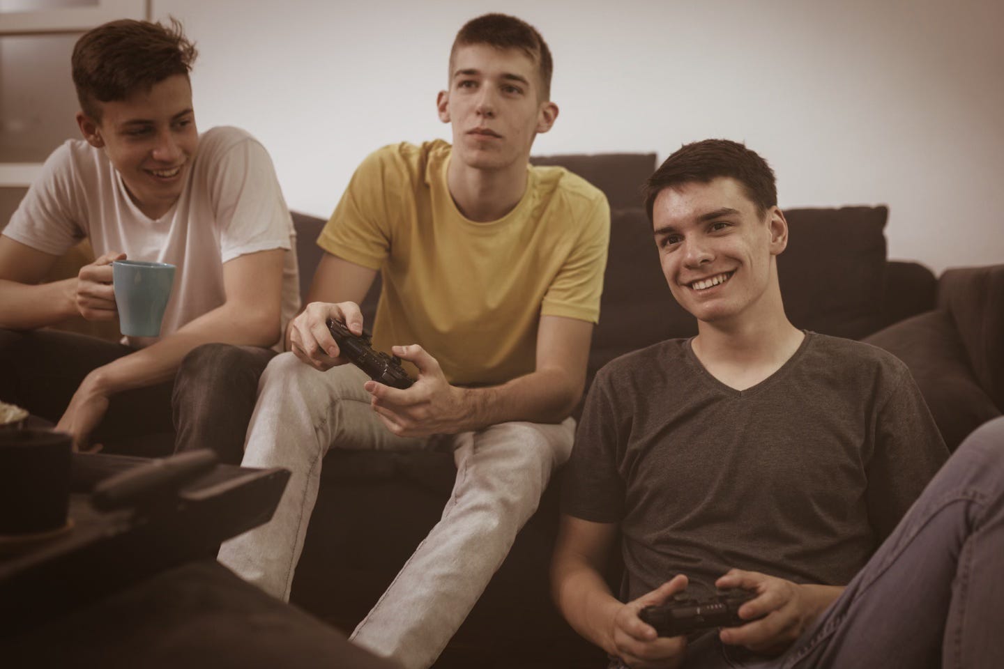 Three teenage boys relax on a couch playing a video game, one holding a controller while another smiles and a third drinks from a mug, all focused on the screen in a casual, comfortable setting.