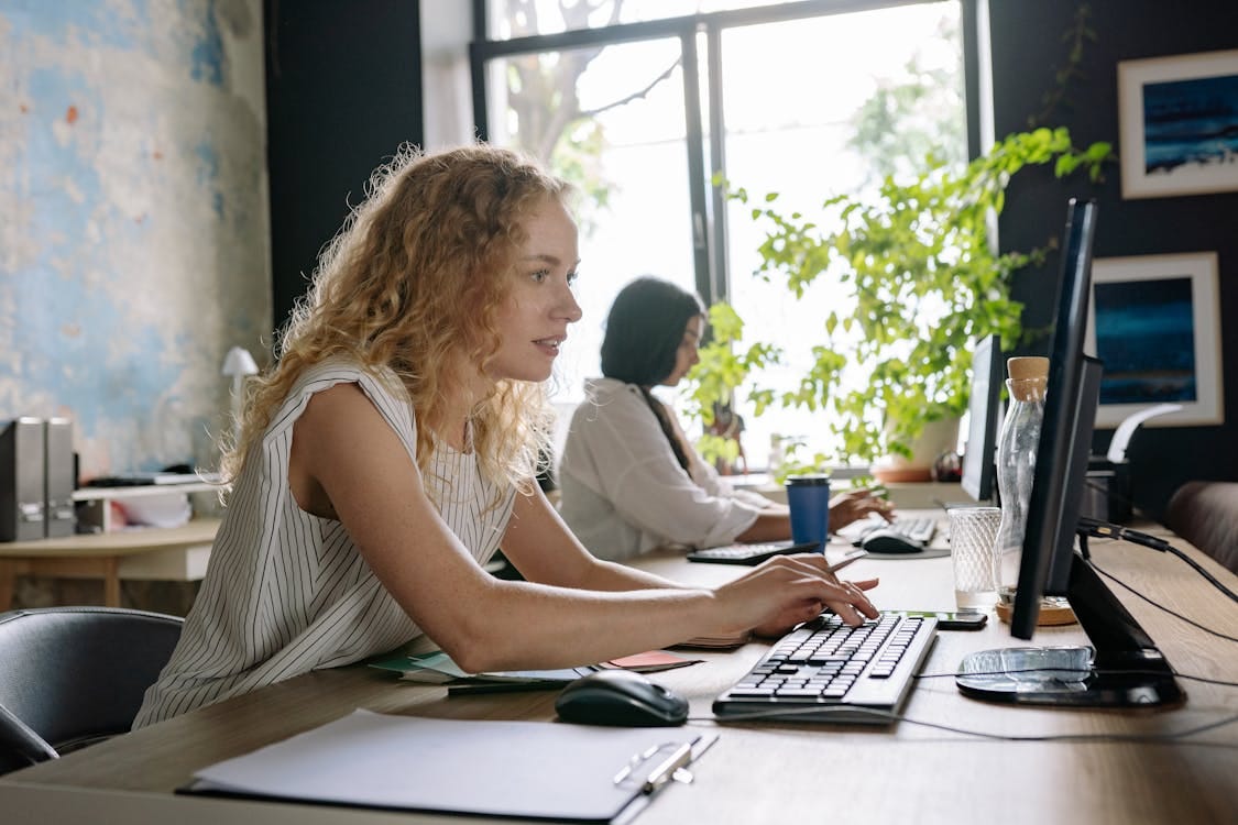 Two focused women working at a bright and modern office space, typing on computers at a shared desk. Stock Photo