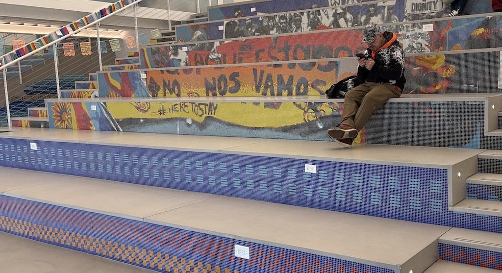 A resident sits on a bench at Make the Road New York, a community center in Corona, Queens, in New York City. Lettering in Spanish reads, "We are here, we're not leaving." The area was one of the largest magnets for asylum-seekers from the border, mostly from Ecuador.