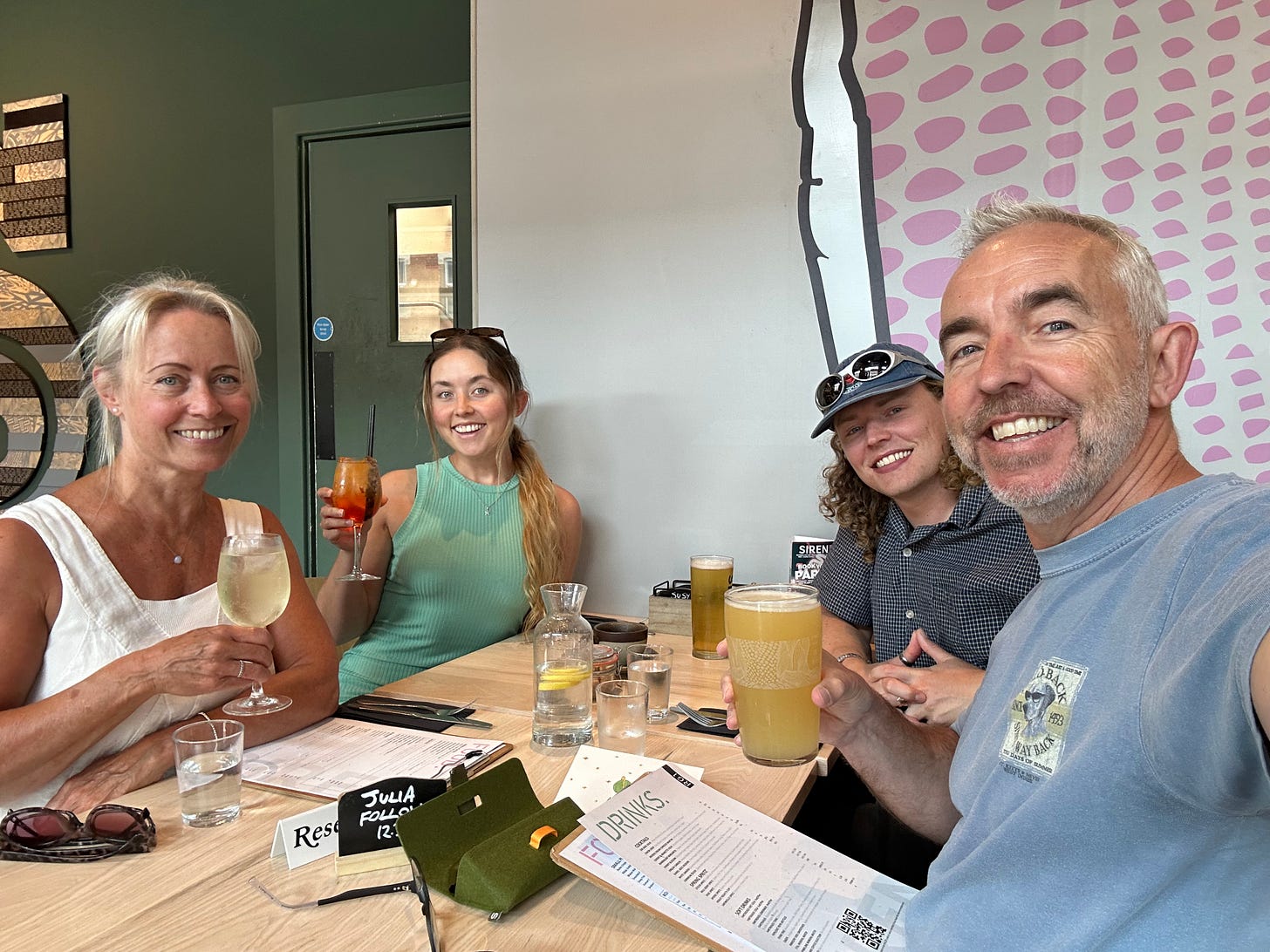 Four smiling family members—two women and two men—sit at a restaurant table raising their drinks in a toast. Menus and a table reservation card labeled “Julia Follows” are visible. Four smiling family members—two women and two men—sit at a restaurant table raising their drinks in a toast. Menus and a table reservation card labeled “Julia Follows” are visible.