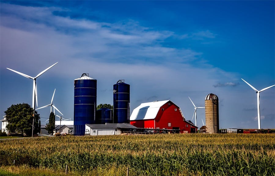 Color photo of a farm with red barn, three bright blue grain silos, an old brown silo, farm buildings, and multiple wind turbines in the near distance Color photo of a farm with red barn, three bright blue grain silos, an old brown silo, farm buildings, and multiple wind turbines in the near distance