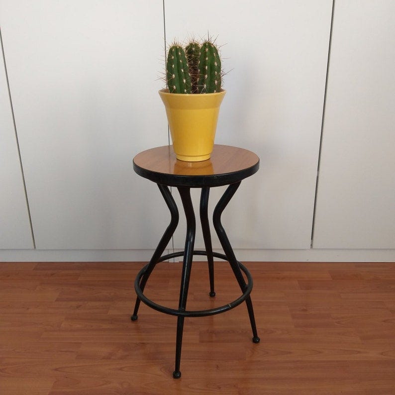 May include: A black metal stool with a round wooden top. A yellow pot with a cactus plant sits on top of the stool.