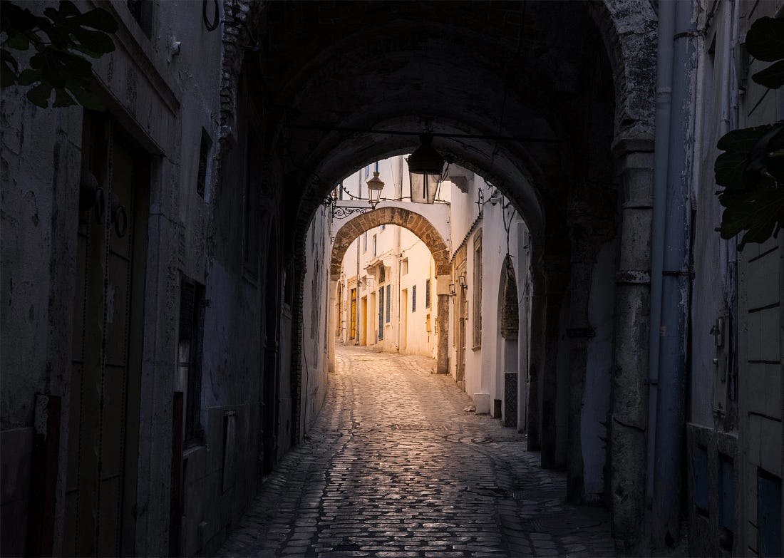 A narrow, arched alley in the Tunis Medina at dusk, where a vaulted passage opens onto softly lit whitewashed buildings and worn stone paving.