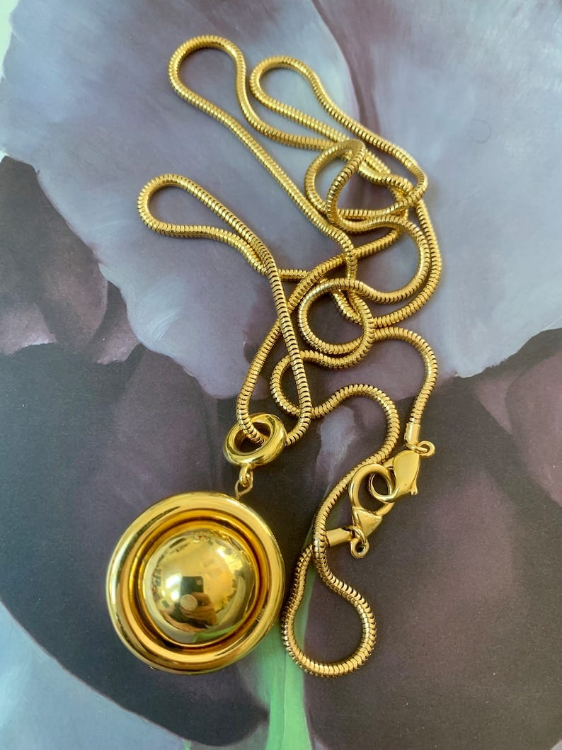 May include: A gold chain necklace with a large round pendant. The pendant has a smooth, shiny surface and is surrounded by a raised, circular rim. The chain is a snake chain style.