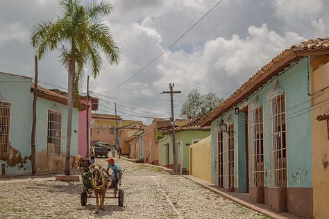 A 3x3 grid of nine photographs depicting life in Cuba. Images include: a boy diving into water; children sitting by a wall; flamingos in a lake; colorful colonial street scenes with classic cars and horse-drawn carts; a cat and a dog in a window; and a lamp-lit cobblestone street at night.
