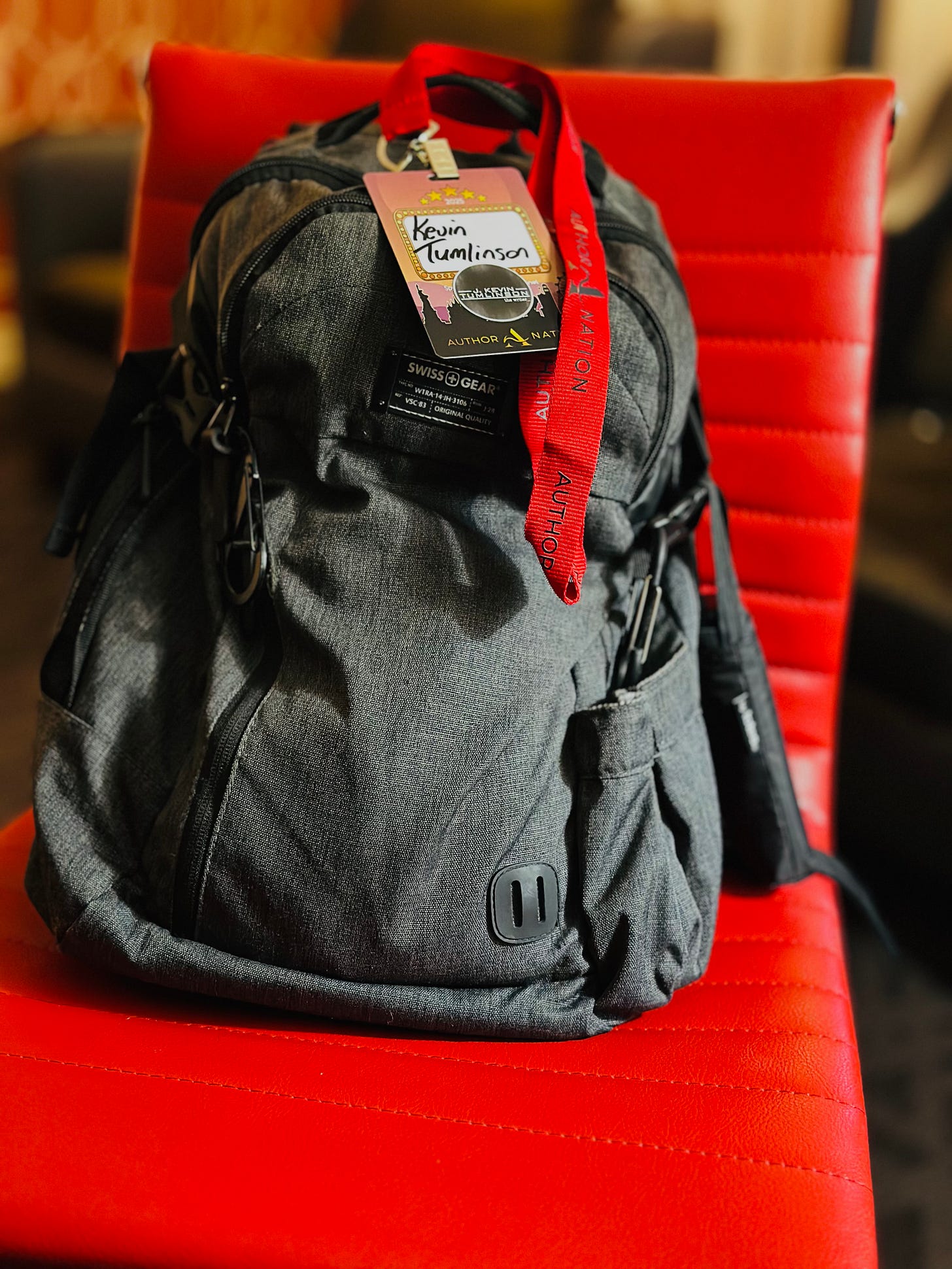 A great backpack resting in an orange desk chair. There is a conference badge with the name “Kevin Tumlinson” written in Sharpie. A red strap is draped over the bag.