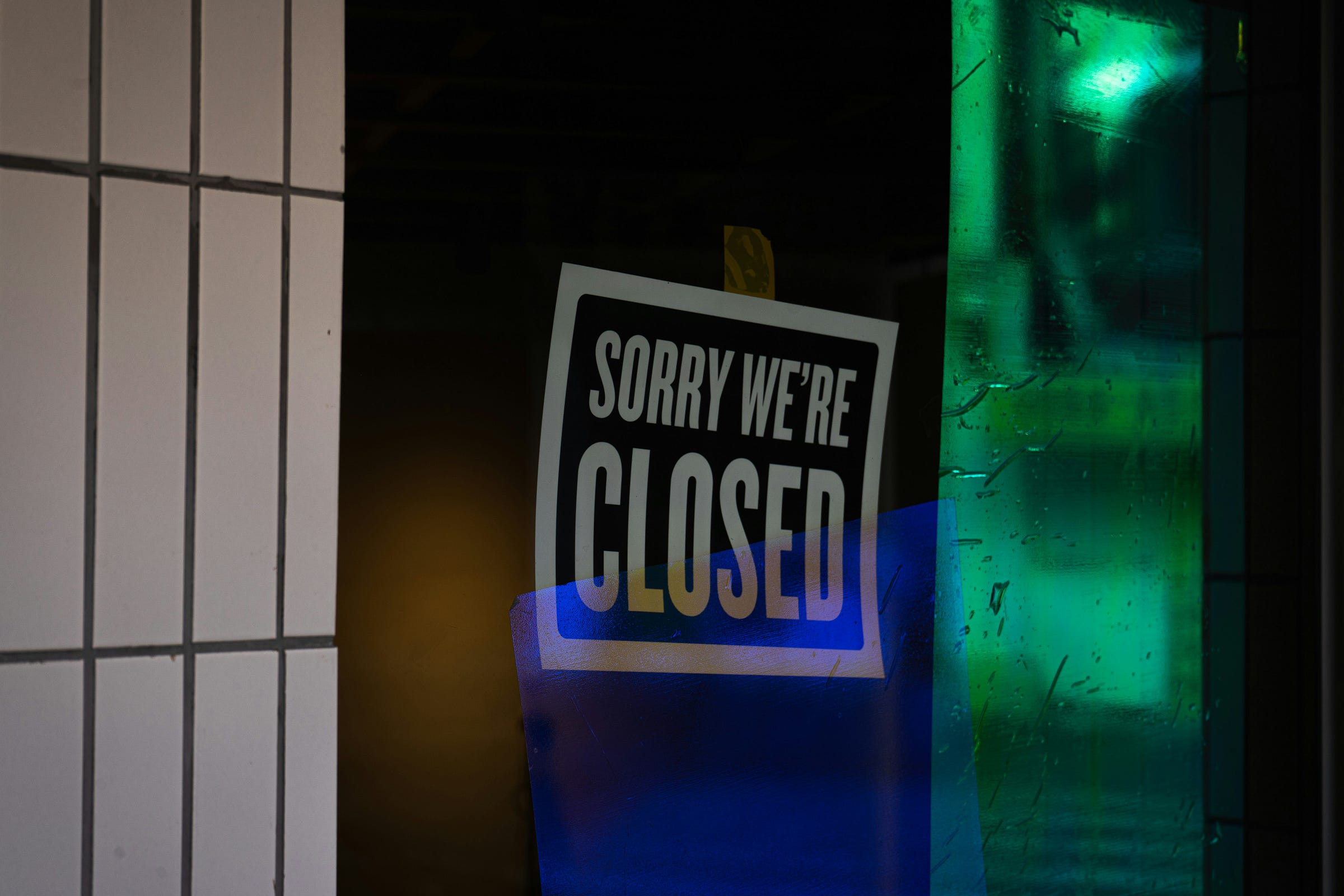 A photograph of a "SORRY WE'RE CLOSED" sign taped to a glass door or window. The sign is black with white text. The surrounding glass reflects a mix of $\textbf{darkness}$, $\textbf{orange}$, $\textbf{blue}$, and $\textbf{bright green}$ light. A portion of a tiled wall is visible on the left.