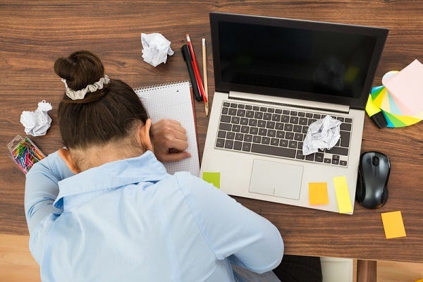 We see someone slumped at a desk, folded over the desktop. Their head rests on their left arm, which is set on a spiral notebook. All around them are crumpled pieces of paper, along with sticky notes and a laptop. The impression is that the person is struggling to write. We see someone slumped at a desk, folded over the desktop. Their head rests on their left arm, which is set on a spiral notebook. All around them are crumpled pieces of paper, along with sticky notes and a laptop. The impression is that the person is struggling to write.