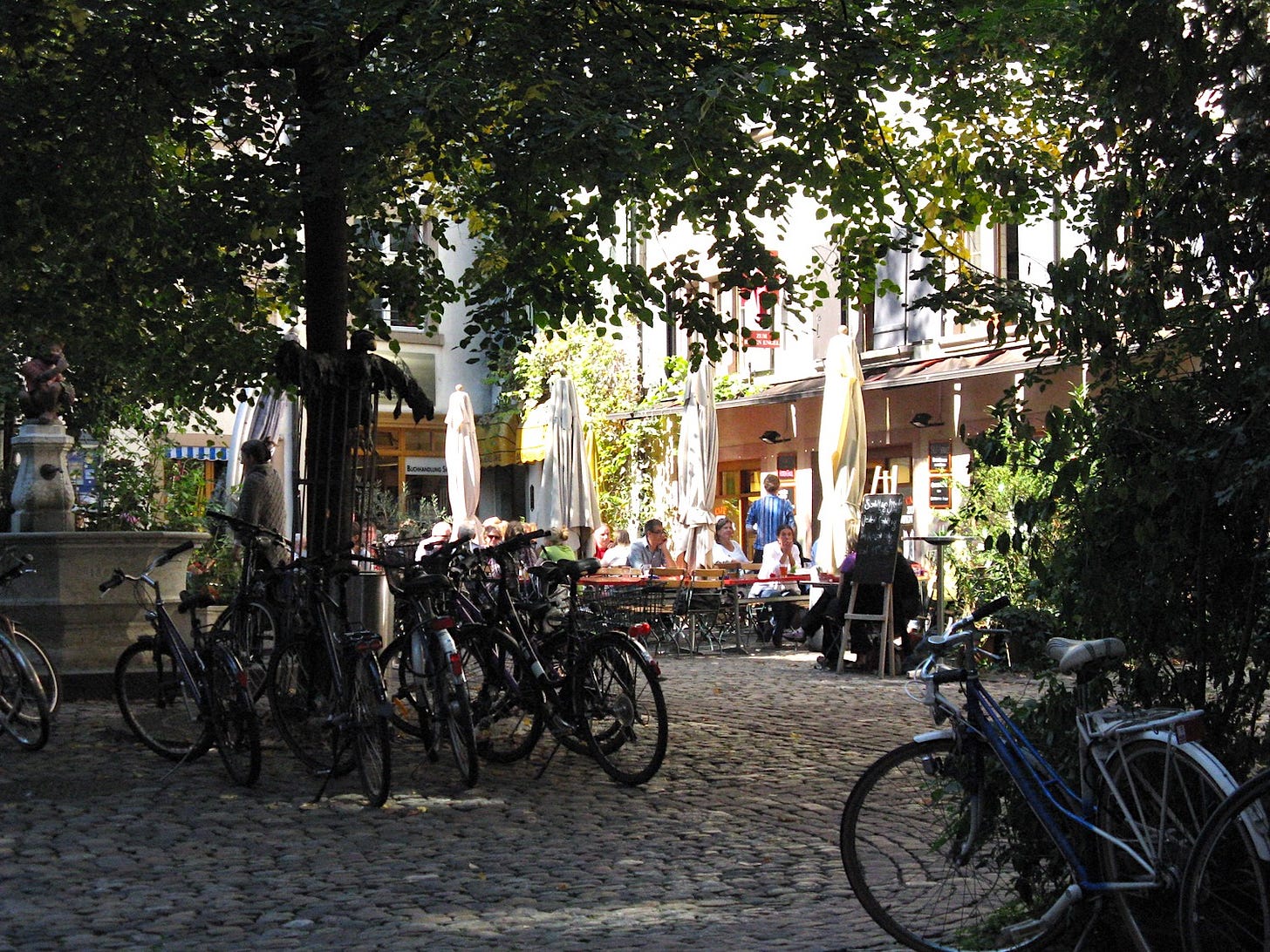 Tree-shaded cobblestone courtyard, parked bicycles, fountain on the left, cafe at the back with outside tables