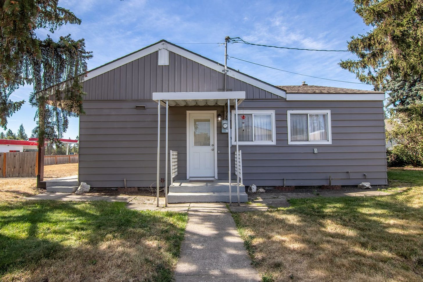 A grey single-storey house with board siding, a small covered entry porch and a concrete path across a plain mowed lawn A grey single-storey house with board siding, a small covered entry porch and a concrete path across a plain mowed lawn