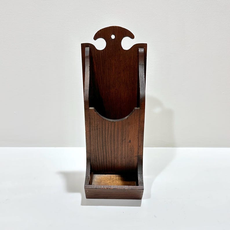 May include: A dark brown wooden wall-mounted match holder. The holder has a rectangular base, a curved front, and a decorative top with a small hole. The wood grain is visible, and the piece appears to be vintage. May include: A dark brown wooden wall-mounted match holder. The holder has a rectangular base, a curved front, and a decorative top with a small hole. The wood grain is visible, and the piece appears to be vintage.