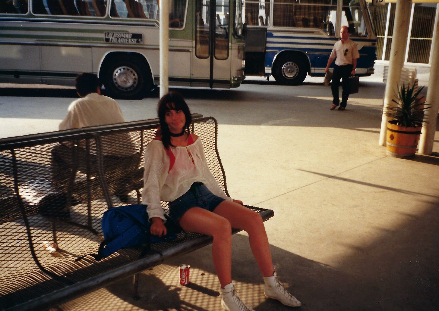 A vintage photograph of a young woman sitting on a metal mesh bench at a bus station. She has dark hair with bangs and is wearing a loose white long-sleeved shirt over a red top, denim shorts, and white high-top sneakers. She has a dark choker necklace. A bright blue bag rests on the bench next to her right hand, and a red Coca Cola can sits on the ground beneath the bench. Behind her, another person is sitting on the opposite side of the bench facing away. In the background, two large buses are parked. One is grey and white with green stripes, and the other is blue and white. A man carrying a briefcase walks across the concrete in the distance. The scene is outdoors under a covered area, with sunlight casting sharp shadows on the ground.