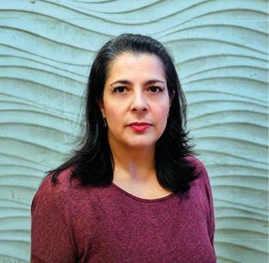 Pessoa com cabelo longo e preto, usando blusa de cor vinho, com maquiagem em tons vermelhos nos lábios, posicionada de frente para a câmera em pé diante de um fundo com padrão de ondas horizontais em tons de azul-claro e cinza. Pessoa com cabelo longo e preto, usando blusa de cor vinho, com maquiagem em tons vermelhos nos lábios, posicionada de frente para a câmera em pé diante de um fundo com padrão de ondas horizontais em tons de azul-claro e cinza.