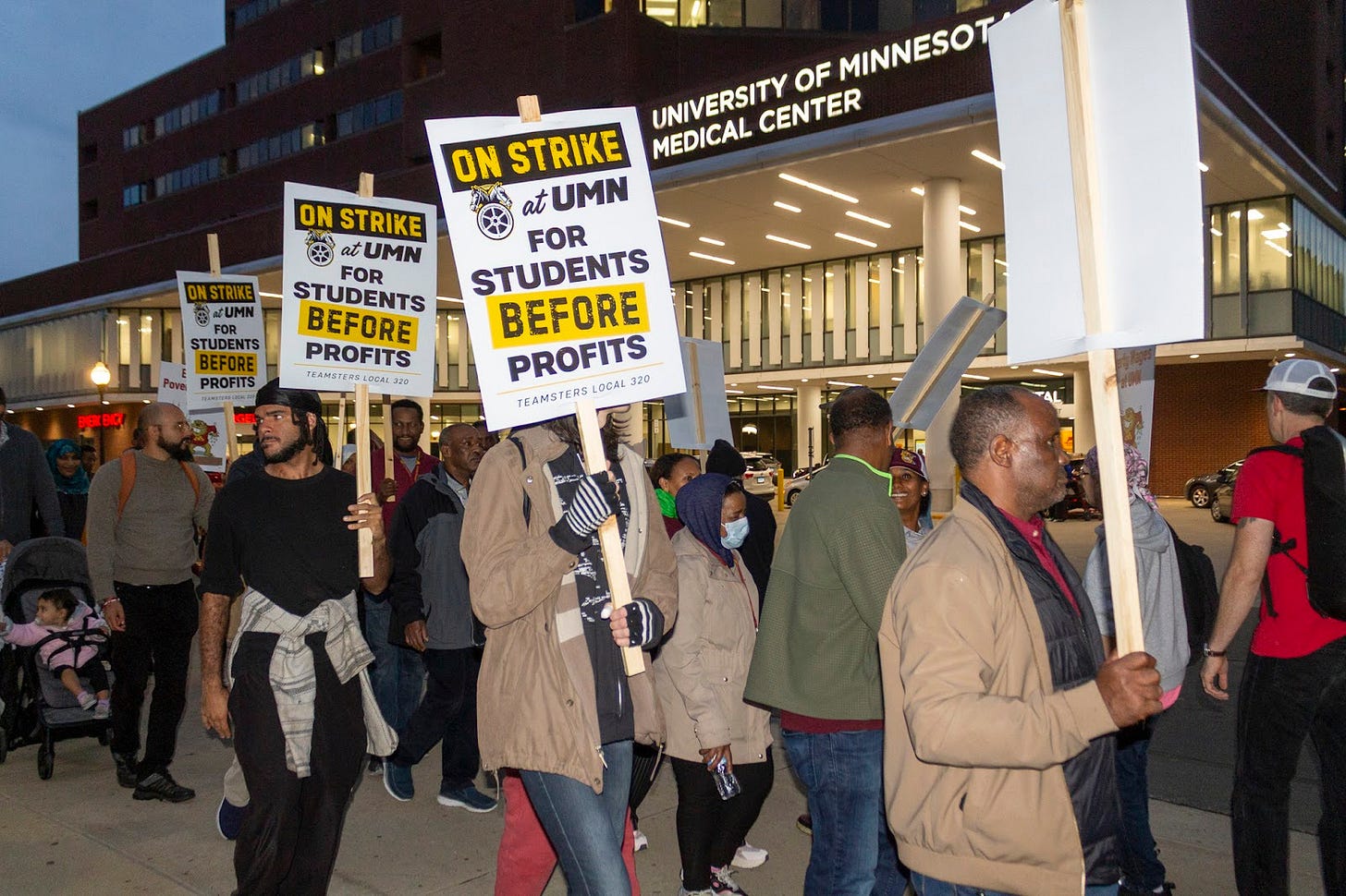 a crowd of people picket in front of the University of Minnesota medical center with signs that read “on strike at UMN for students before profits"
