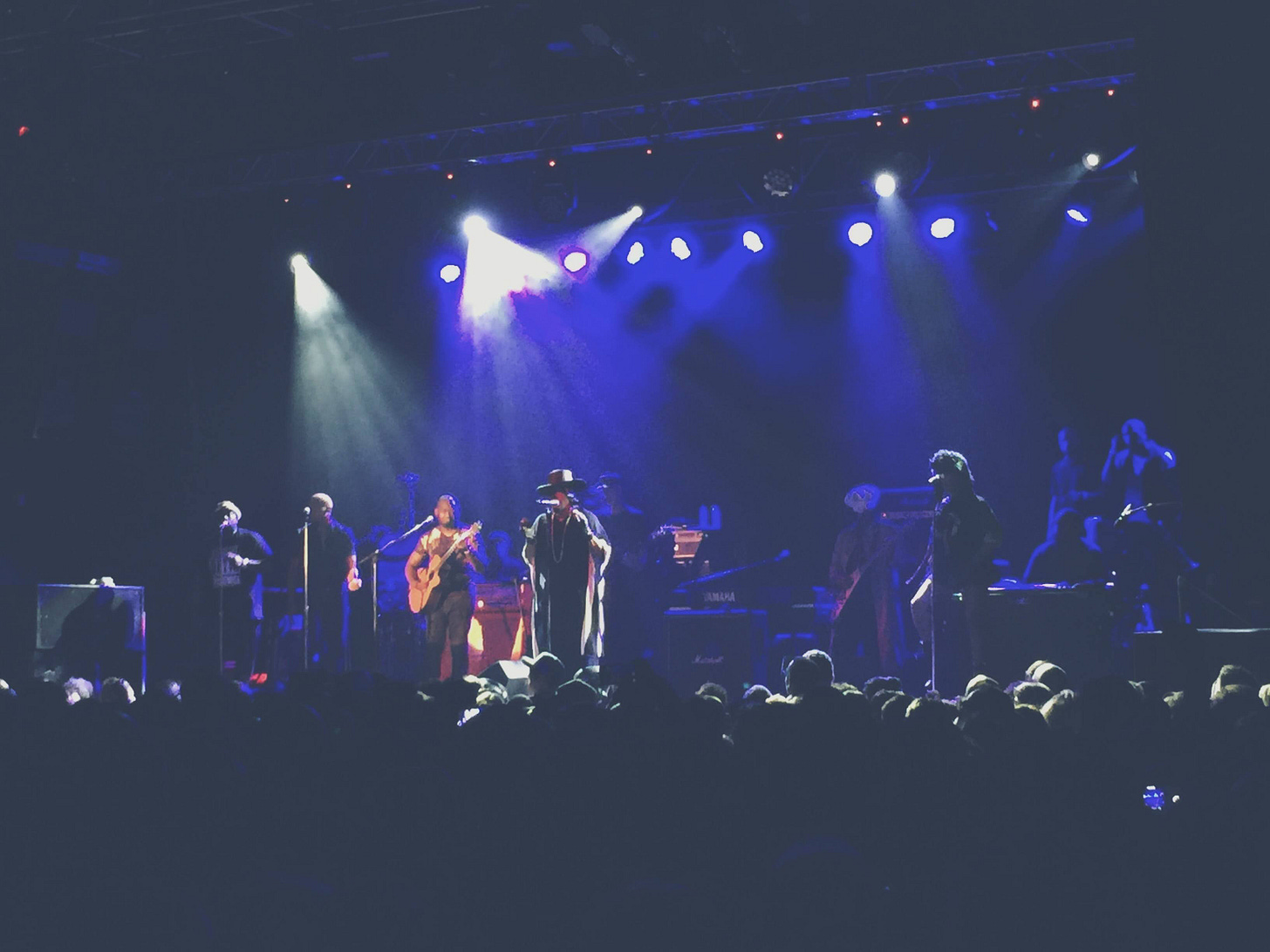 A wide-angle shot of a live concert stage, showing D'Angelo and the Vanguard performing under bright stage lights with a blue and purple hue and in front of a large audience. The crowd is silhouetted in darkness as the Vanguard play guitars, sing, and move under the bright, theatrical lighting . A wide-angle shot of a live concert stage, showing D'Angelo and the Vanguard performing under bright stage lights with a blue and purple hue and in front of a large audience. The crowd is silhouetted in darkness as the Vanguard play guitars, sing, and move under the bright, theatrical lighting .
