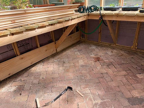 Alt text: Three side-by-side photos of a greenhouse interior during construction. The images show newly built wooden raised planting beds arranged around the perimeter of the space, with a herringbone-pattern brick floor installed throughout the center walkway. The greenhouse framing and clear wall panels are visible, and the beds are unfinished, showing the structural supports beneath the bench tops. The photos capture different angles of the same space, highlighting the layout of the raised beds and brick flooring.