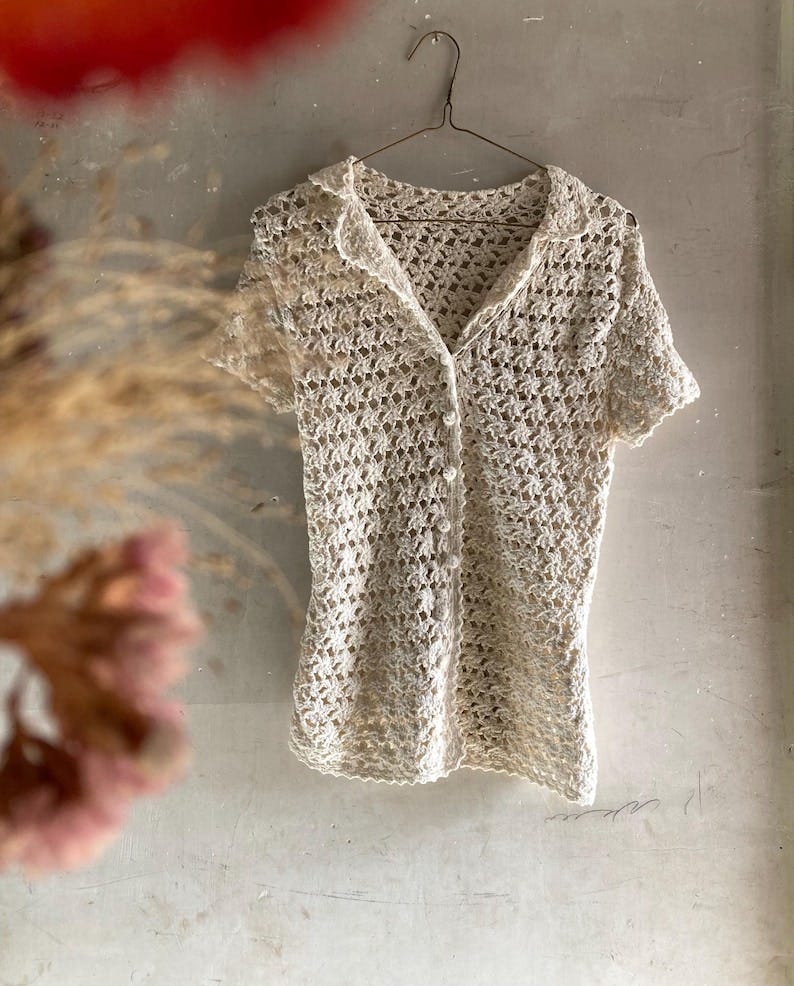 May include: A beige crocheted short-sleeved button-up top hangs on a dark metal hanger against a textured light beige wall. The top has a collared neckline and a delicate, open-weave pattern. The garment is lightweight and airy, suitable for warm weather.  The crocheted fabric is evenly textured and the buttons are small and unobtrusive. The overall style is simple and elegant.