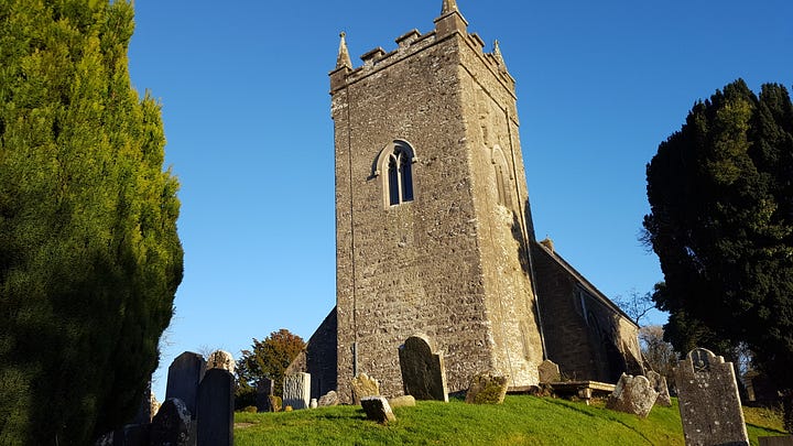 1. old font and standing stone in the graveyard. 2. The medieval tower. 3. Close-up of arched window high on tower wall with carved figure slightly above and to the left. 4. Zoom capture of the stone head with very weathered body barely visible.