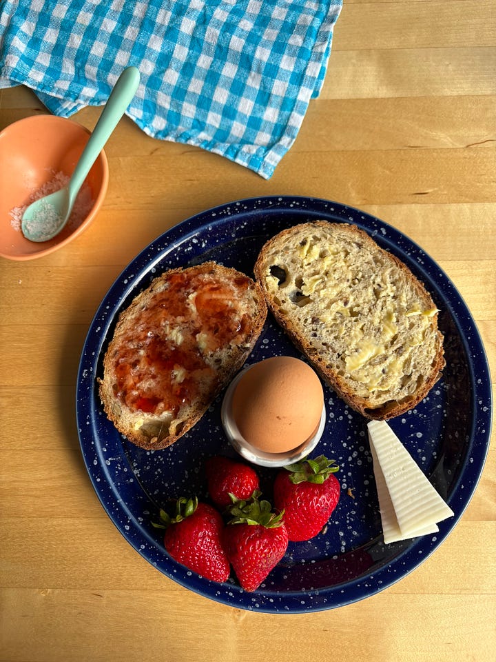 Two breakfast plates featuring a boiled egg, toast with jam, fresh fruit (watermelon in one, strawberries in the other) and pieces of cheese (cheddar in one, goats cheese in the other)