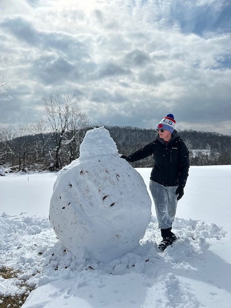 Woman in a Bills beanie and black jacket holds a small snowball, and then a basketball-sized snowball, and then stands next to a giant snowball that is almost as tall as her.