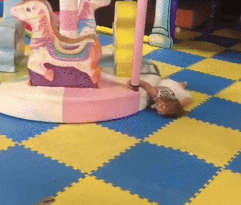 A giph of a little girl being dragged face down in a circle indoors by a small merry go round.