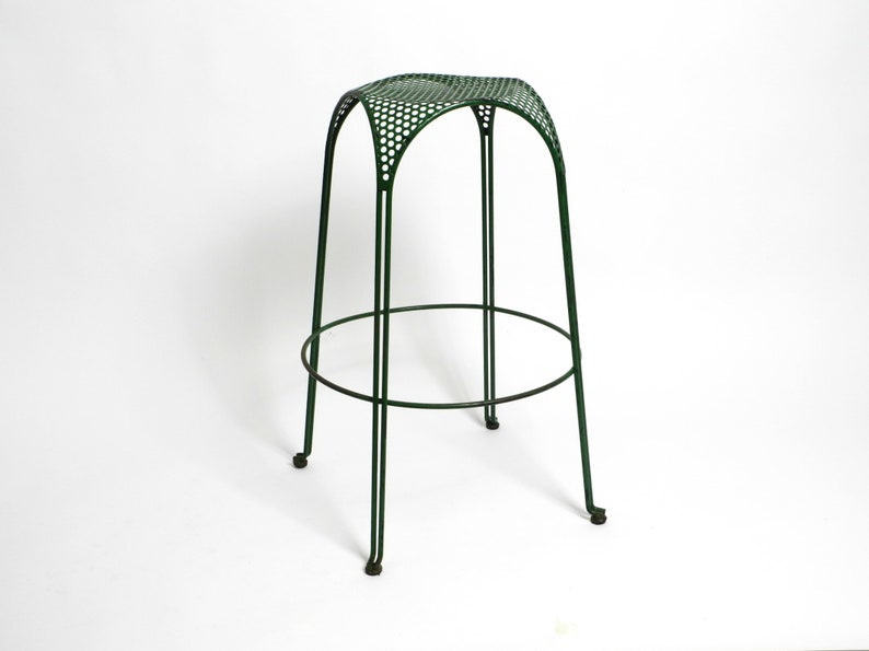 May include: A dark green metal bar stool with a perforated seat and a circular footrest. The stool has a modern, minimalist design with slender legs and a slightly arched back. The metal appears to have a smooth finish. The stool is shown against a plain white background.