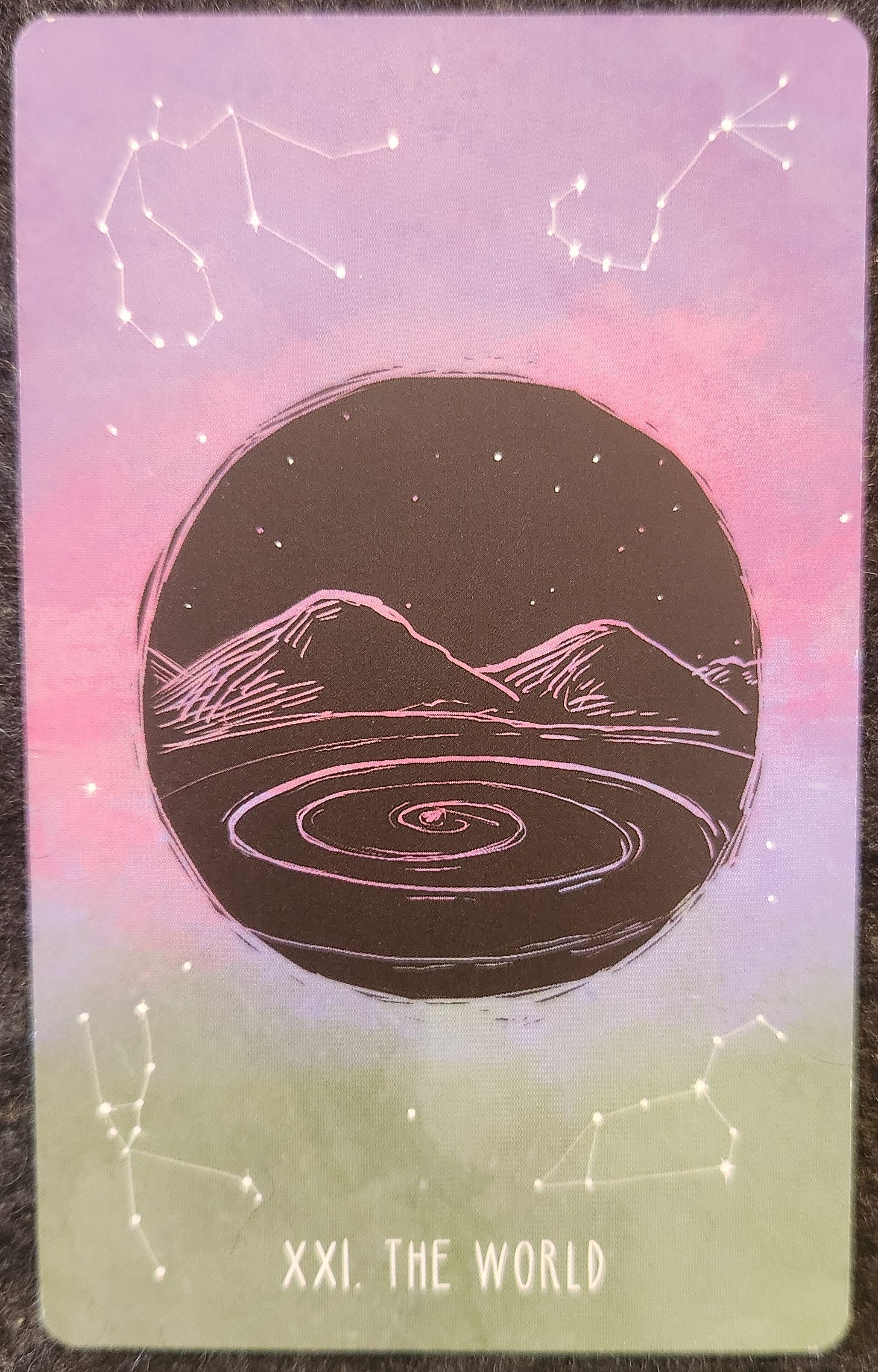 A tarot card labelled XXI. The World. A black circle with a lake and mountain scene etched into it with white ink. The lake shows a swirling vortex on the surface. Behind the black circle is a pink and green sky with constellations of stars in each corner of the card. 