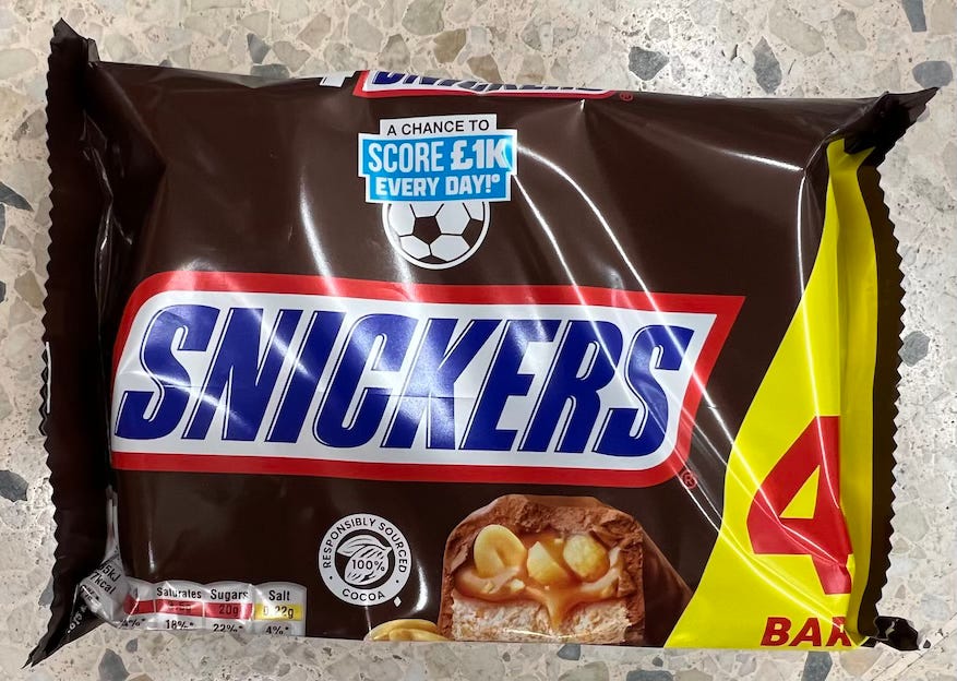 Image of a Snickers chocolate bar multipack containing four bars. The packaging is mostly dark brown with the SNICKERS logo prominently displayed in blue and white with a red outline. A football-themed promotion near the top reads “A CHANCE TO SCORE £1K EVERY DAY!” The bottom left shows nutritional information, and the bottom right has a close-up image of the bar’s cross-section, revealing caramel, nougat, and peanuts. A yellow panel on the right displays the number “4” and the word “BARS.”