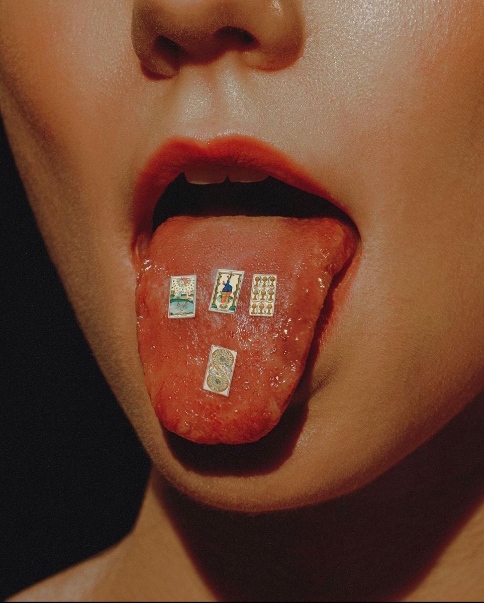 Close-up of a woman’s mouth with tongue extended displaying miniature tarot cards, dramatic beauty photography with warm lighting.