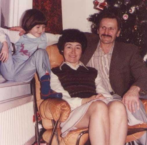 A family photo from the late 1970s of a 5 year old girl in pyjamas, sitting with her Mum and Dad who are both seated and smiling at the camera. Mother and daughter both have 1970s bowl cuts and father has a 1970s handlebar moustache. There is a Christmas tree behind the group.