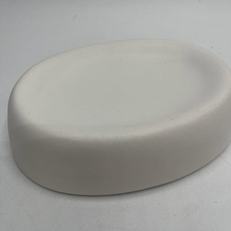 May include: A white ceramic soap dish with a rounded oval shape and a smooth surface.