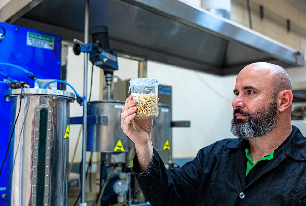 Pessoa com barba e cabeça raspada, vestindo jaqueta preta com gola verde fluorescente, segura com a mão direita um recipiente transparente contendo grãos de milho estourado, em ambiente industrial com equipamentos azuis ao fundo, tanques metálicos e avisos de segurança amarelos com símbolo de triângulo. Pessoa com barba e cabeça raspada, vestindo jaqueta preta com gola verde fluorescente, segura com a mão direita um recipiente transparente contendo grãos de milho estourado, em ambiente industrial com equipamentos azuis ao fundo, tanques metálicos e avisos de segurança amarelos com símbolo de triângulo.