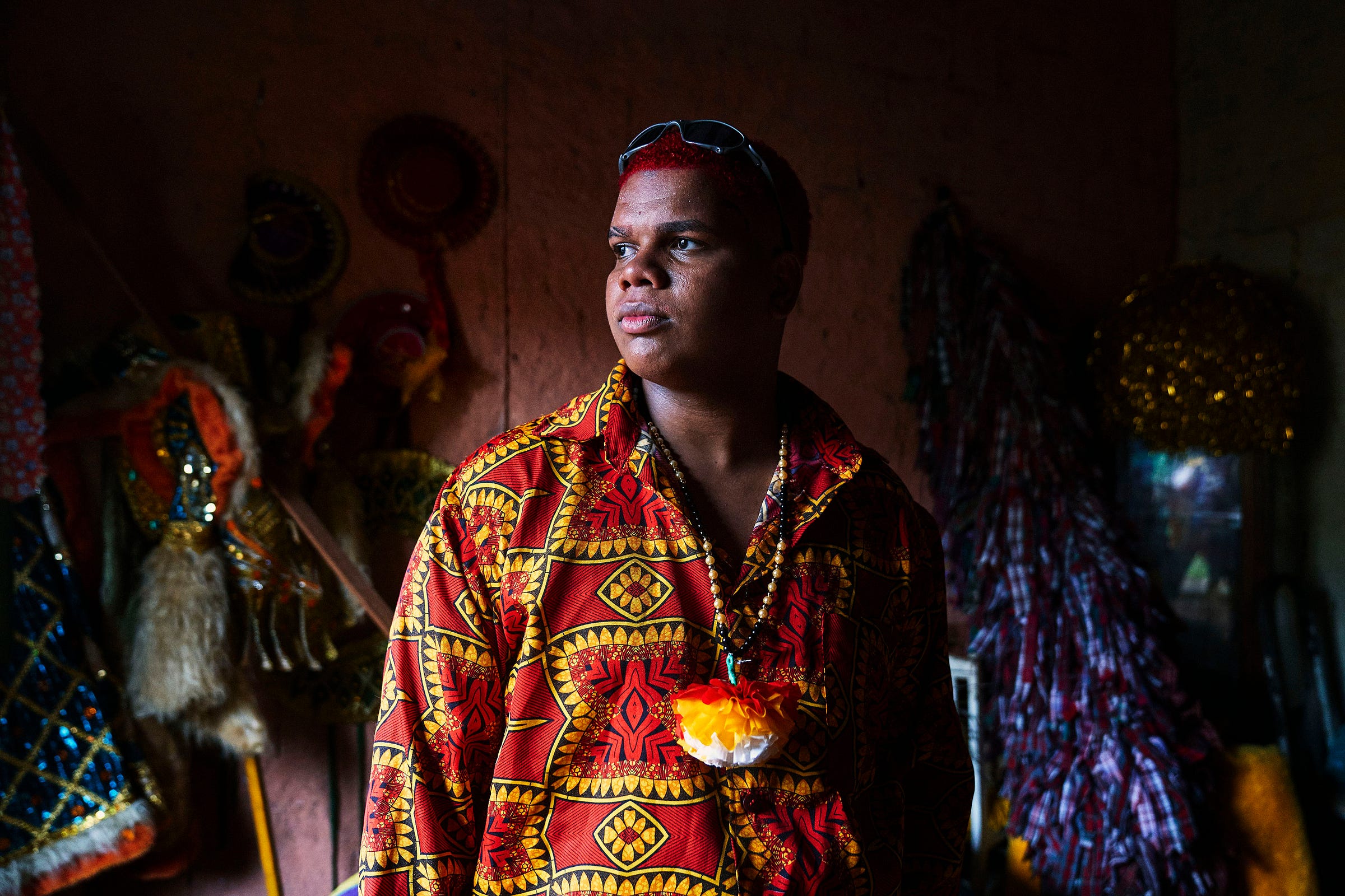 A person with short red hair and glasses atop their head wears a colorful red and yellow patterned shirt, standing indoors with traditional costumes and decorations in the dim background.