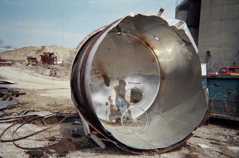 a color photo of a bicycle wheel standing on its own inside some kind of metal cylinder with the diameter of a tall person. around it lie various metal detritus and behind it in the distance is a dirt hill.