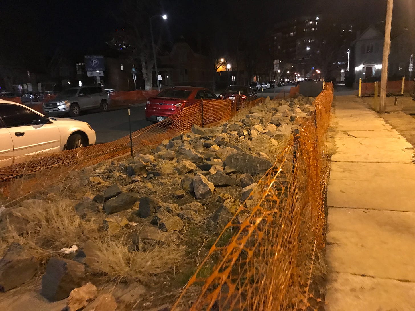A picture of a boulevard at night. The boulevard, between the sidewalk and curb, is filled with small to medium sized boulders. It is fenced off with orange plastic fencing anchored on green rebar posts.
