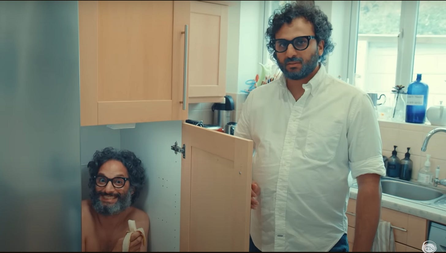 A mostly naked Jason Mantzoukas crouches in a cupboard, smiling and eating a banana. Nish Kumar stands next to the cabinet, dressed like Mantzoukas.
