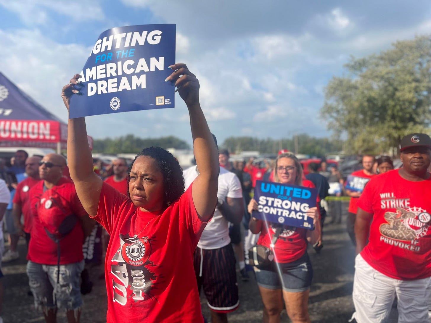 A Black woman wearing a red shirt stands in front of other people wearing red UAW shirts holding blue posters reading "Fighting for the American dream" A Black woman wearing a red shirt stands in front of other people wearing red UAW shirts holding blue posters reading "Fighting for the American dream"