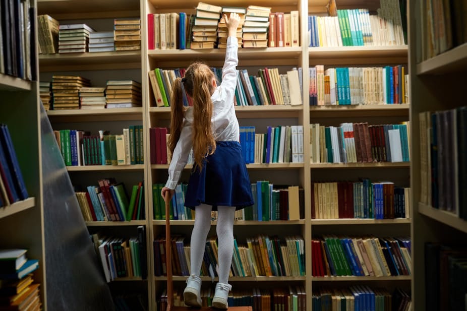 #descrevemospravocê  A imagem mostra uma menina em uma biblioteca, tentando alcançar um livro em uma das prateleiras mais altas. Ela está de costas, em pé sobre um banquinho de madeira. Com o braço direito totalmente estendido para cima, ela tenta pegar um dos livros posicionados na fileira superior da estante. Na mão esquerda, ela segura uma haste fina de madeira, possivelmente usada como apoio para alcançar livros.  A menina tem cabelos longos e castanhos, presos em um rabo de cavalo com um laço rosa. Ela veste uma camisa branca de mangas compridas, uma saia azul-marinho, meia-calça branca e sapatos claros. A biblioteca está repleta de estantes de madeira clara, lotadas de livros organizados em fileiras horizontais, com capas de diversas cores, principalmente tons de verde, azul, vermelho e bege. A iluminação é suave, com destaque voltado para a estante central, onde a menina está.  A cena transmite um ambiente acolhedor, de curiosidade e busca pelo conhecimento, com foco na autonomia da criança dentro do espaço da biblioteca.