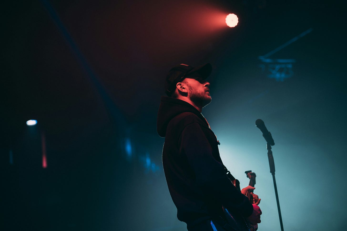 A man in a black hoodie stands on a stage in front of a microphone holding a guitar.