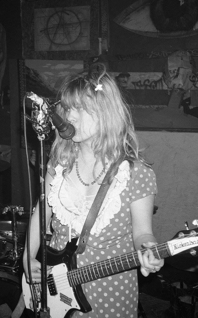 A black and white photo of Kat Bjelland of Babes in Toyland in a spotty dress playing guitar and singing into the mic