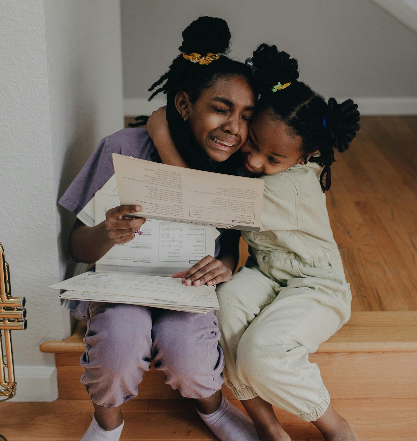 Two young siblings hugging each other, as one of them cries and holds up a piece of paper.