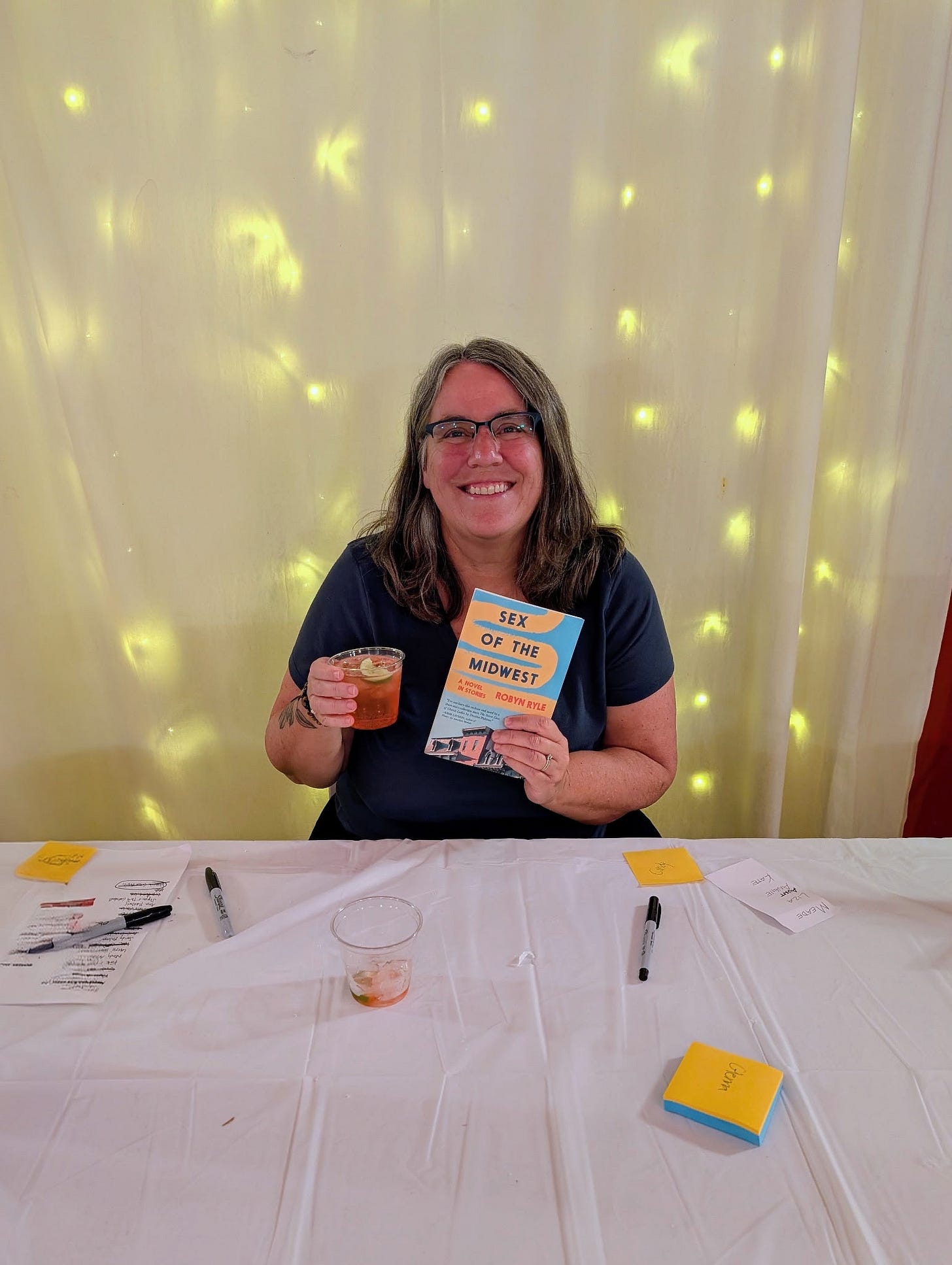 Robyn sitting in front of twinkle lights, grinning widely and holding a copy of SEX OF THE MIDWEST and a cocktail