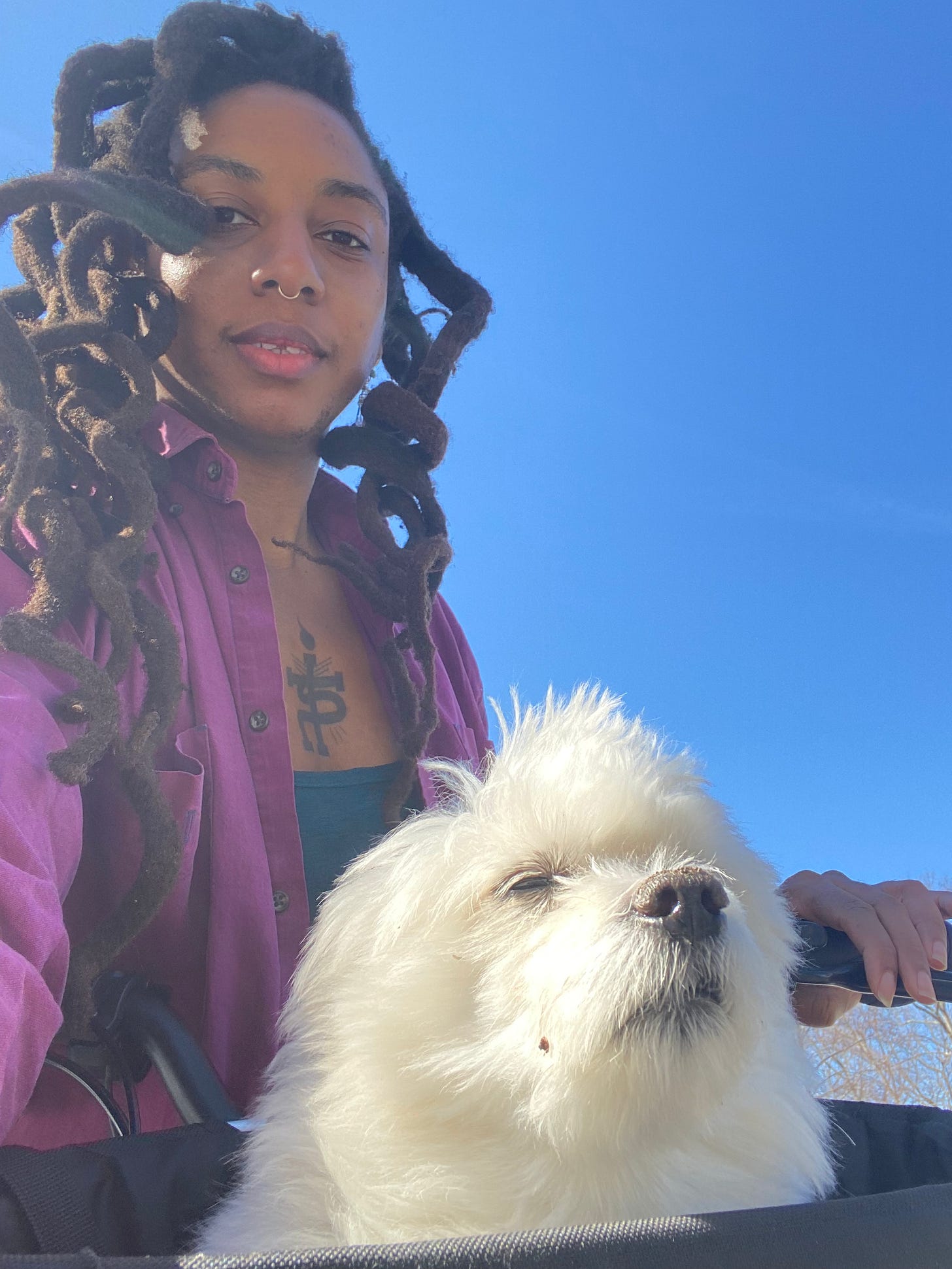 beautiful black woman with locs smiling on her bicycle. A white pomerian dog sits in her bike basket, gazing forward