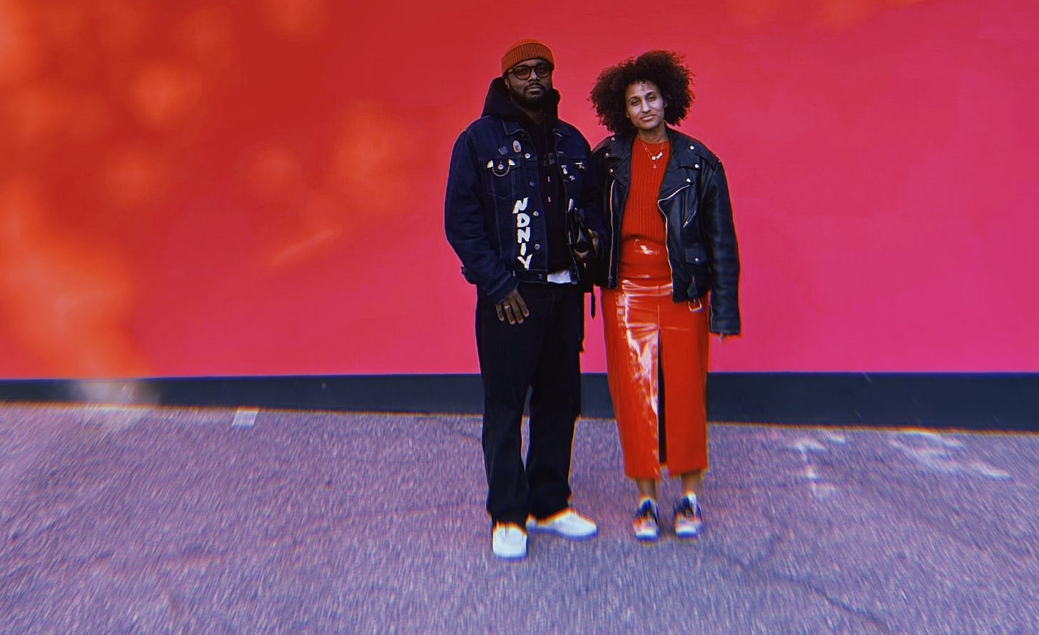 ​Dr. Cadet stands in front of a red background wearing a red oversized knit sweater, pleather red skit, vintage black leather biker jacket, and silver sneakers. Her natural curls are full and her facial expression is serious just like Robert, a Black man standing next to her who wears a red knit beanie, sunglasses, a black hoodie, dark jean jacket adorned with his art, black jeans and white sneakers. They are here to disrupt the art game.