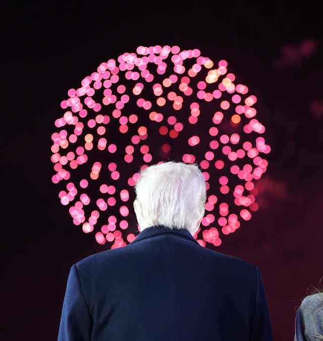 A white-haired man and a woman with dark-blond hair have their backs to the camera as they look at pink and red fireworks. A white-haired man and a woman with dark-blond hair have their backs to the camera as they look at pink and red fireworks.