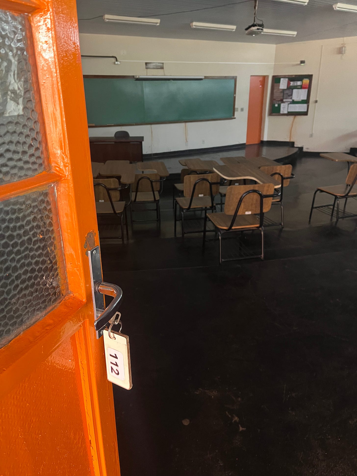 Foto colorida na vertical. Mostra partde uma porta de sala de aula com a chave na fechadura com um chaveiro com o númer 112. Ao fundo, carteiras do modelo universitário de madeira e um quadro negro