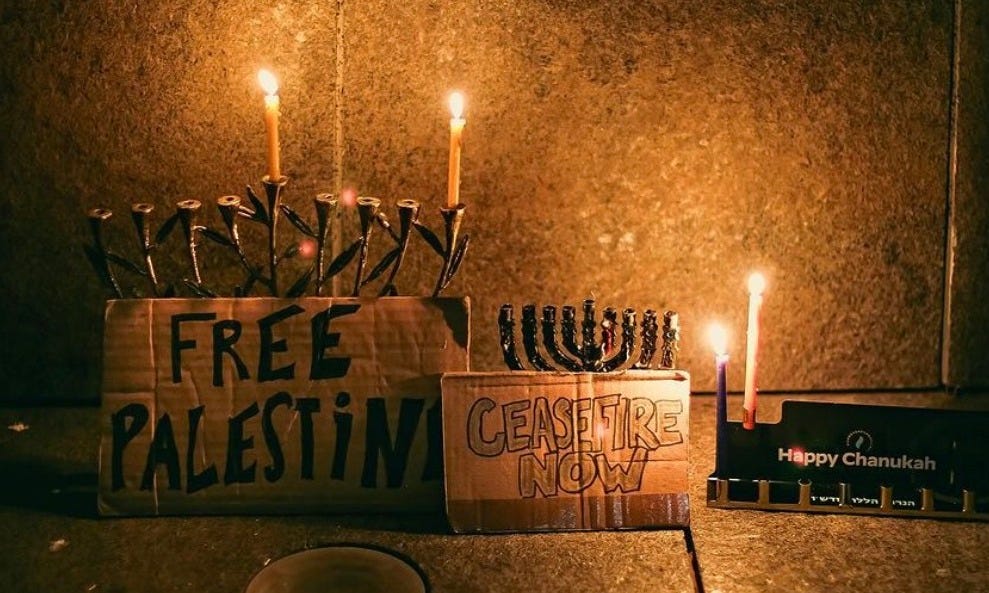 street altar with Hanukkah menorahs and signs reading "free Palestine" and "Ceasefire Now"... the light is dim and candles burn brightly