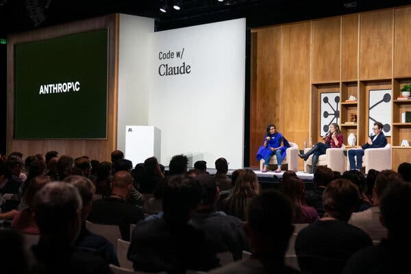 Three people sit to the right in round seats on a stage, with an audience in the foreground. To the left, the words “Anthropic” and “Code w/Claude” appear on large panels.   