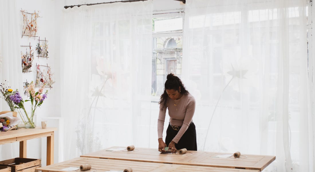 A woman standing in a white and airy studio with flowers on the wall, leaning on a wooden table with twine balls on top of it